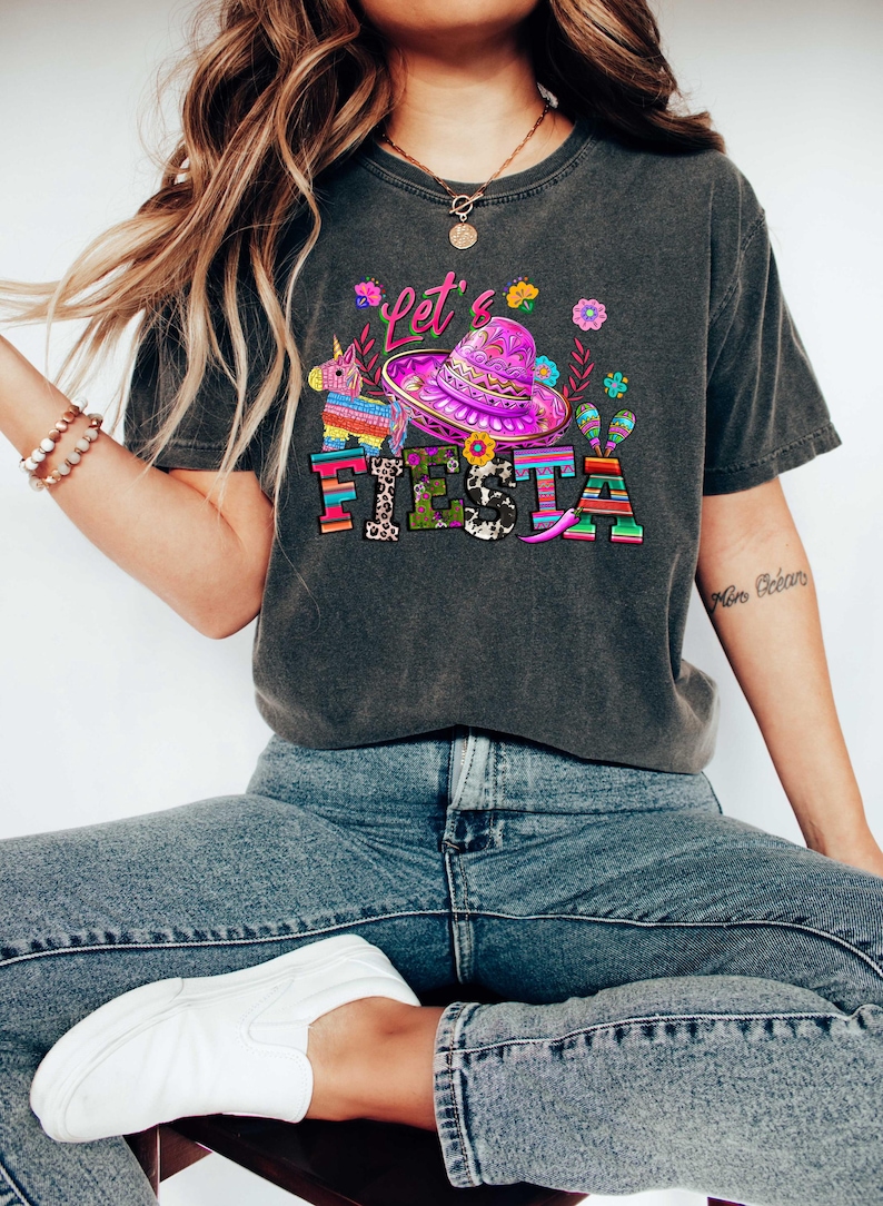 May include: Dark gray t-shirt with a colorful "Let's Fiesta" graphic. The design includes a sombrero, a pi&ntilde;ata, maracas, and floral elements. The shirt is a casual style, suitable for festive occasions.