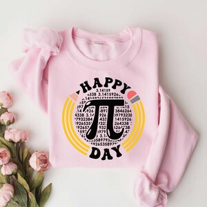 May include: A pink sweatshirt with a graphic design of a large black pi symbol surrounded by yellow and orange pencils. The text "Happy Pi Day" is written above and below the pi symbol. The pi symbol is surrounded by numbers.