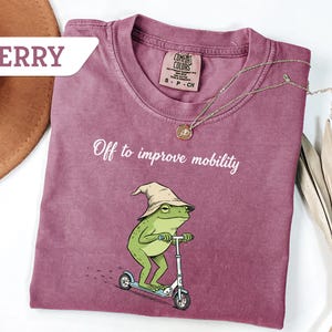 May include: Berry-colored t-shirt with the text "Off to improve mobility" and a cartoon frog riding a scooter. The frog wears a hat. A gold necklace is draped across the shirt. The shirt has a Comfort Colors tag.