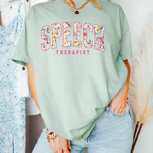 May include: A light green t-shirt with a floral print design that spells out "SPEECH THERAPIST" in pink letters.
