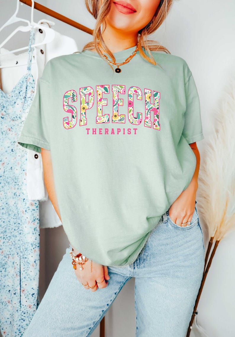 May include: A light green t-shirt with a floral print design that spells out "SPEECH THERAPIST" in pink letters.