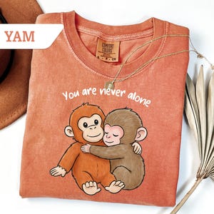 May include: A coral-colored t-shirt featuring a graphic of two hugging cartoon monkeys with the text "You are never alone." A gold necklace is draped over the shirt. The shirt is folded and has a tag that says "Comfort Colors."