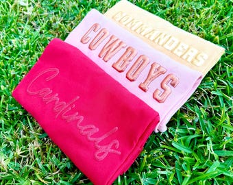 May include: Three folded t-shirts stacked diagonally. The top shirt is tan with the word COMMANDERS in block letters. The middle shirt is light pink with the word COWBOYS in block letters. The bottom shirt is red with the word Cardinals in script.