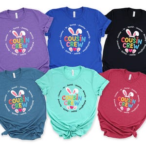 May include: Six short-sleeved t-shirts in shades of purple, blue, black, teal, green, and red. Each shirt displays a circular graphic with the words "COUSIN CREW" and bunny ears, with names encircling the design.