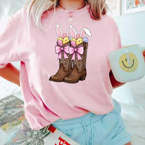 May include: Pink t-shirt with a graphic of two brown cowboy boots filled with Easter eggs and bunny ears. The boots have pink bows.