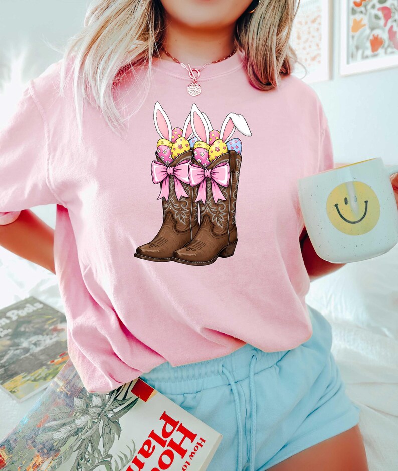 May include: Pink t-shirt with a graphic of two brown cowboy boots filled with Easter eggs and bunny ears. The boots have pink bows.