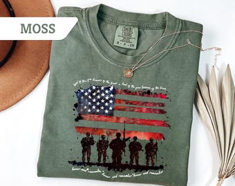 Puede incluir: Una camiseta verde con un diseño de bandera americana en acuarela y siluetas de soldados. El texto "land of the free because of the brave - land of the free because of the brave - honor and remember - honor and remember - honor and remember" está impreso en la camiseta.