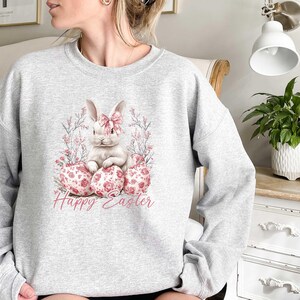 May include: A gray sweatshirt with a white bunny rabbit sitting on pink and white Easter eggs. The bunny has a pink bow and is surrounded by pink flowers. The text "Happy Easter" is printed below the bunny.