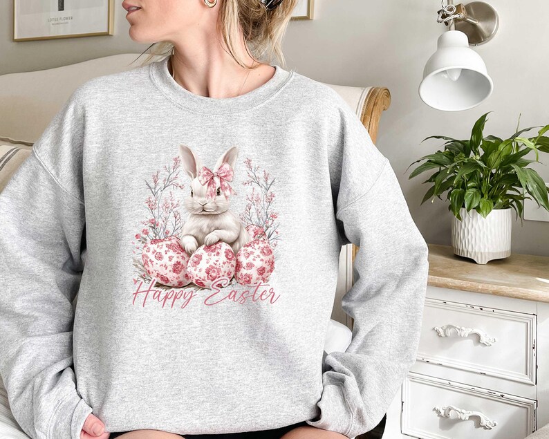 May include: A gray sweatshirt with a white bunny rabbit sitting on pink and white Easter eggs. The bunny has a pink bow and is surrounded by pink flowers. The text "Happy Easter" is printed below the bunny.