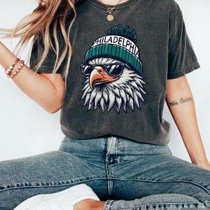 Puede incluir: Una camiseta gris oscuro con un gr&aacute;fico de un &aacute;guila calva con una boina verde y blanca con la palabra "PHILADELPHIA" en ella. El &aacute;guila lleva gafas de sol.