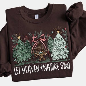 May include: A brown sweatshirt with a Christmas-themed graphic. The design features three stylized Christmas trees, a pink bow, and the text "LET HEAVEN & NATURE SING" in white. The trees are decorated with stars and berries.