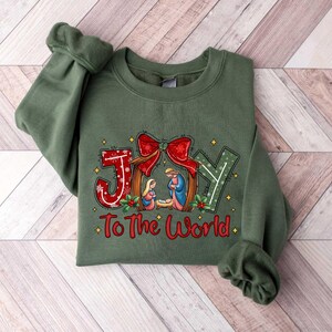 May include: Green sweatshirt with a Christmas-themed design featuring the word "JOY" with a nativity scene and the text "To The World".