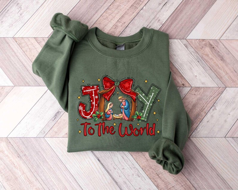 May include: Green sweatshirt with a Christmas-themed design featuring the word "JOY" with a nativity scene and the text "To The World".