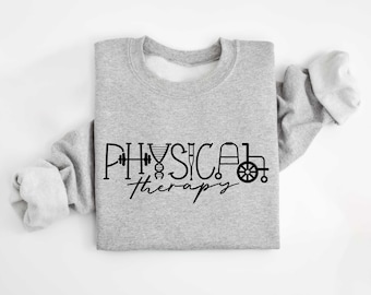 May include: A pale grey sweatshirt featuring the words "Physical Therapy" in black script, accompanied by themed icons. These include a dumbbell, DNA strand, crutch, walker, and wheelchair. The sweatshirt is displayed on a white background.