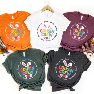 May include: Five t-shirts in various colours, including orange, white, burgundy, dark green, and charcoal grey. Each shirt has a circular design with the words "Cousin Crew", bunny ears, and hearts. Names are printed around the circle.