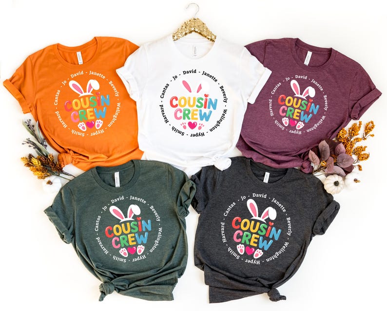 May include: Five t-shirts in various colours, including orange, white, burgundy, dark green, and charcoal grey. Each shirt has a circular design with the words "Cousin Crew", bunny ears, and hearts. Names are printed around the circle.
