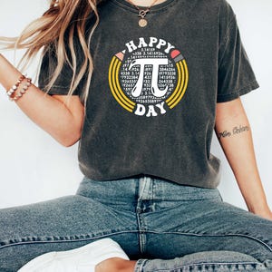 May include: A dark gray t-shirt with a graphic of a circle with the mathematical symbol pi and the text "HAPPY PI DAY" in a retro font. The circle is surrounded by yellow and orange stripes.