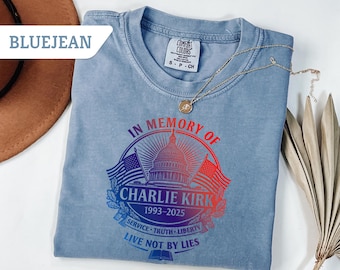 May include: Bluejean Comfort Colors t-shirt with a graphic that reads "IN MEMORY OF CHARLIE KIRK 1993-2025" with the US Capitol building and flags. The shirt also has the phrases "SERVICE TRUTH LIBERTY" and "LIVE NOT BY LIES". A gold necklace is draped across the shirt.