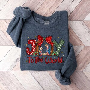 May include: A dark blue sweatshirt with a Christmas-themed design. The design features a manger scene with the words "Joy to the World" printed below.