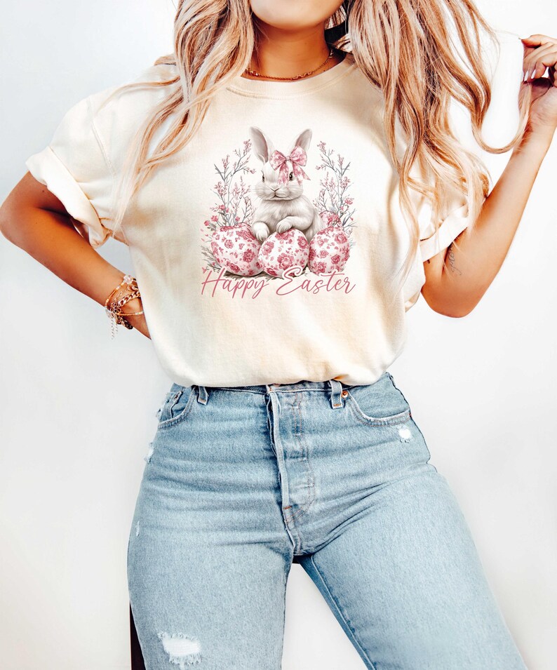 May include: A white t-shirt with a watercolor illustration of a white bunny rabbit wearing a pink bow and sitting on three pink and white Easter eggs. The text "Happy Easter" is printed below the bunny.