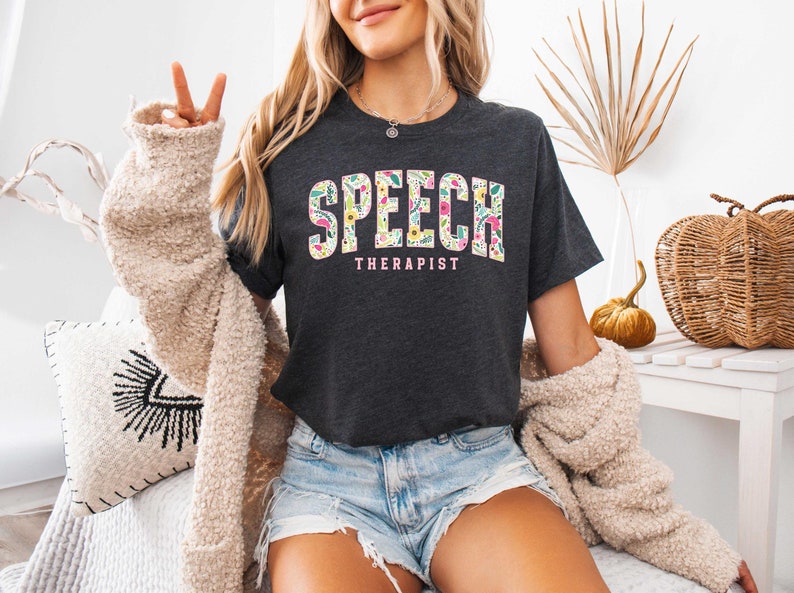 May include: A dark grey t-shirt with a floral design spelling out "SPEECH THERAPIST" in pink letters.
