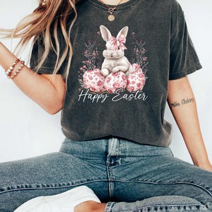 May include: A dark gray t-shirt with a white bunny rabbit wearing a pink bow sitting on pink and white floral Easter eggs. The text "Happy Easter" is printed below the bunny.