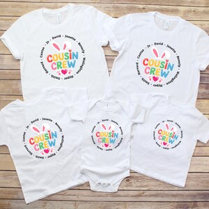 May include: A collection of white t-shirts and a babygrow featuring a colourful "Cousin Crew" design. The design includes bunny ears, hearts, and names. The items are displayed on a wooden surface.