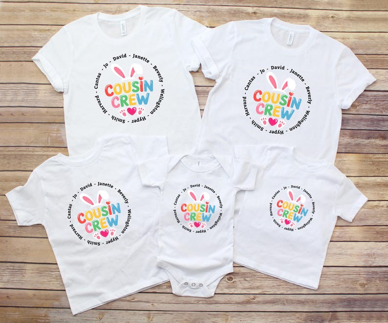 May include: A collection of white t-shirts and a babygrow featuring a colourful "Cousin Crew" design. The design includes bunny ears, hearts, and names. The items are displayed on a wooden surface.