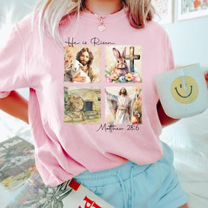 May include: Pink t-shirt with a graphic design of a religious scene. The design includes a lamb, a cross, a bunny, and a man with a halo. The text "He is Risen" and "Matthew 28:6" are included in the design.