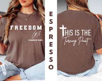 May include: Espresso-colored t-shirt with the word "FREEDOM" in white block letters on the front, along with a signature and the name "Charlie Kirk." The back of the shirt features a white cross and the phrase "THIS IS THE Turning Point."