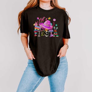 May include: Black t-shirt with a colorful graphic that says "Let's Fiesta". The graphic includes a sombrero, flowers, a unicorn pi&ntilde;ata, and the word "Fiesta" in bright colors. The shirt is a casual style.