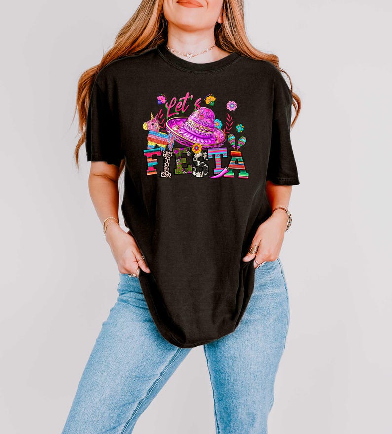 May include: Black t-shirt with a colorful graphic that says "Let's Fiesta". The graphic includes a sombrero, flowers, a unicorn pi&ntilde;ata, and the word "Fiesta" in bright colors. The shirt is a casual style.
