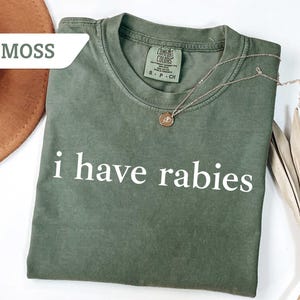Może przedstawiać: Zielony t-shirt w kolorze mchu z nadrukiem "i have rabies" w kolorze białym na przodzie. Koszulka wykonana jest z miękkiego, wygodnego materiału i ma luźny kr&oacute;j.