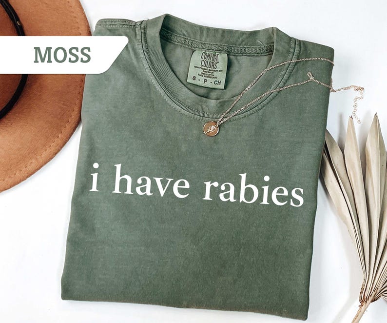 Może przedstawiać: Zielony t-shirt w kolorze mchu z nadrukiem "i have rabies" w kolorze białym na przodzie. Koszulka wykonana jest z miękkiego, wygodnego materiału i ma luźny kr&oacute;j.