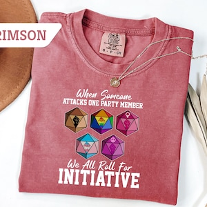 May include: Crimson-colored t-shirt with the text "When Someone Attacks One Party Member, We All Roll For Initiative." The shirt features colorful geometric dice designs, and a Comfort Colors tag.