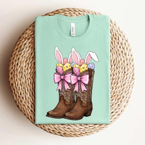May include: A light green t-shirt with a graphic of two brown cowboy boots filled with colorful Easter eggs and bunny ears. The boots have pink bows and are on a light brown wicker background.