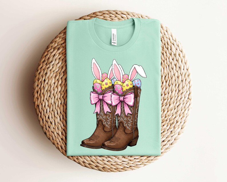 May include: A light green t-shirt with a graphic of two brown cowboy boots filled with colorful Easter eggs and bunny ears. The boots have pink bows and are on a light brown wicker background.