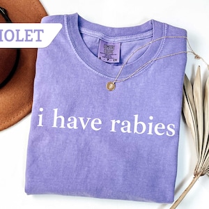 Może przedstawiać: Fioletowy T-shirt z nadrukiem "i have rabies" w kolorze białym na przodzie. Koszulka wykonana jest z miękkiego, wygodnego materiału.