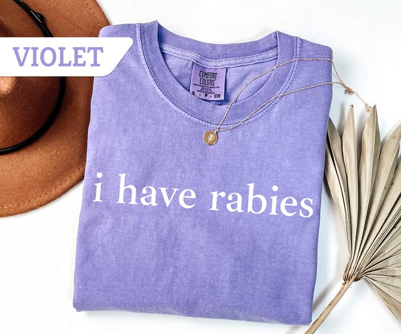 Może przedstawiać: Fioletowy T-shirt z nadrukiem "i have rabies" w kolorze białym na przodzie. Koszulka wykonana jest z miękkiego, wygodnego materiału.