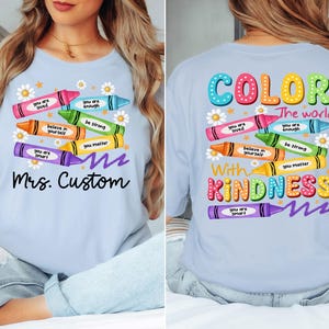K&ouml;nnte beinhalten: Hellblaues T-Shirt mit bunten Buntstift-Grafiken und positiven Botschaften. Auf der Vorderseite steht "Mrs. Custom", auf der R&uuml;ckseite "Color the world with kindness".