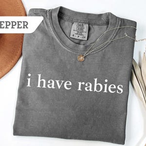 Może przedstawiać: Szary T-shirt z nadrukiem "i have rabies" w kolorze białym. Koszulka wykonana jest z miękkiego, wygodnego materiału i ma luźny kr&oacute;j.