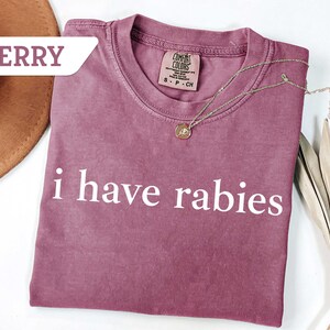 Może przedstawiać: Koszulka w kolorze jagodowym z nadrukiem "i have rabies" w kolorze białym.