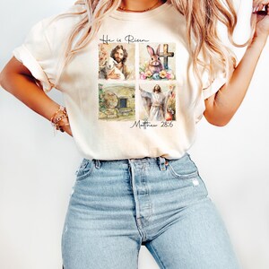 May include: A light beige t-shirt with a watercolor design of four images. The images are of a lamb, a cross, a rabbit, and a man with his arms raised. The text "He is Risen" and "Matthew 28:6" are printed on the shirt.