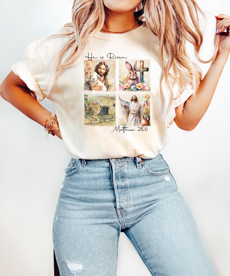 May include: A light beige t-shirt with a watercolor design of four images. The images are of a lamb, a cross, a rabbit, and a man with his arms raised. The text "He is Risen" and "Matthew 28:6" are printed on the shirt.
