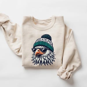 Puede incluir: Una sudadera beige con un gr&aacute;fico de un &aacute;guila que lleva una boina verde y blanca con la palabra "PHILADELPHIA" en ella. El &aacute;guila lleva gafas de sol.