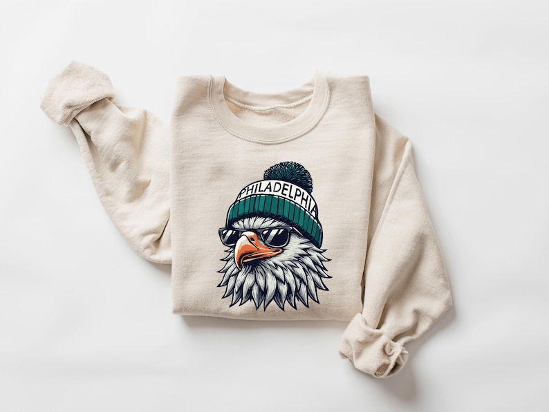 Puede incluir: Una sudadera beige con un gr&aacute;fico de un &aacute;guila que lleva una boina verde y blanca con la palabra "PHILADELPHIA" en ella. El &aacute;guila lleva gafas de sol.