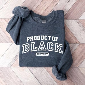 Puede incluir: Una sudadera gris oscura con el texto "PRODUCT OF BLACK HISTORY" en letras blancas.