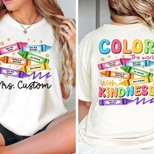 K&ouml;nnte beinhalten: Cremefarbenes T-Shirt mit einem Buntstift-Design und positiven Botschaften. Auf der Vorderseite steht "Mrs. Custom", auf der R&uuml;ckseite "Color the world with kindness". Das T-Shirt hat einen Rundhalsausschnitt und kurze &Auml;rmel.