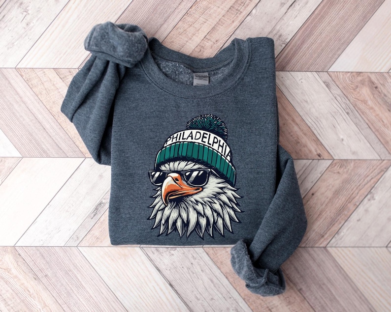 Puede incluir: Una sudadera gris oscuro con un gr&aacute;fico de un &aacute;guila que lleva una boina verde y blanca con la palabra "PHILADELPHIA" en ella. El &aacute;guila lleva gafas de sol.