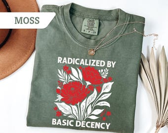 May include: Moss green t-shirt with the words "RADICALIZED BY BASIC DECENCY" in white. The shirt features a floral design with red flowers and white leaves. A gold necklace with a pendant is draped on the shirt.